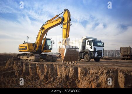 Riesiger gelber Raupenbagger und ein Baukipper, der daneben steht, während er an einem sonnigen Tag gegen einen blauen Himmel arbeitet. Betonung des großen b Stockfoto