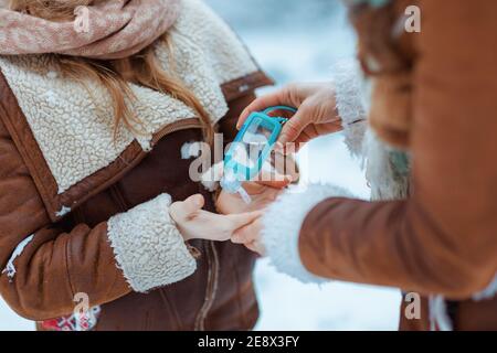 Nahaufnahme von Mutter und Tochter in Hüten und Schaffellmänteln Desinfektion Hände mit Desinfektionsmittel im Freien im Stadtpark im Winter. Stockfoto
