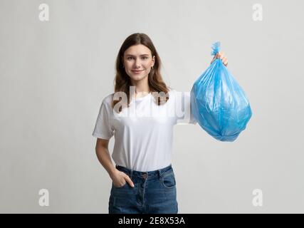 Glückliche junge Frau hält blauen Plastikmüllbeutel, auf grauem Hintergrund stehend Stockfoto