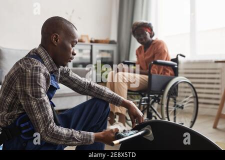 Seitenansicht Porträt von afroamerikanischen Mann Montage Möbel in Wohnräumen mit Frau im Rollstuhl im Hintergrund, Handwerker-Service und Assistan