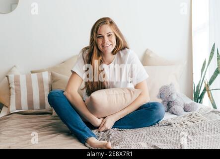 Die rothaarige Frau mit Sommersprossen ist entspannt und lachend in einem schlichten weißen T-Shirt zu Hause auf dem Bett mit einem Kissen. Stockfoto