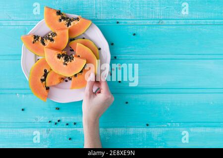 Hand mit Papaya-Scheibe auf blauem Hintergrund. Teller mit Papaya-Stücken. Speicherplatz kopieren. Draufsicht. Stockfoto