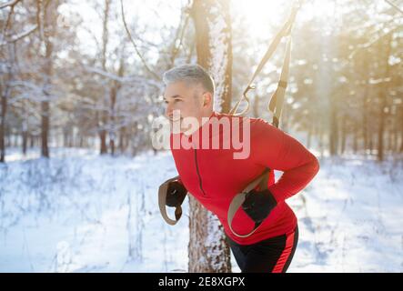 Starker reifer Mann, der im verschneiten Winterpark TRX Fitness Workout macht. Ausdauerübungen im Freien Stockfoto