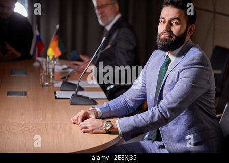 arabischer Mann in Anzug sitzt am Schreibtisch posiert Blick auf die Kamera, gut lächelnde männliche Executive zufrieden mit guten Job Karriere posiert in modernen Sitzungssaal Stockfoto