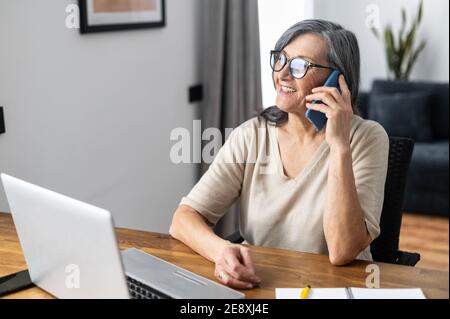 Beschäftigte ältere Frau mittleren Alters trägt eine Brille mit einem Laptop für Remote-Arbeit von zu Hause aus reden auf dem Smartphone und lächelt. Fröhliches Senior Business Stockfoto