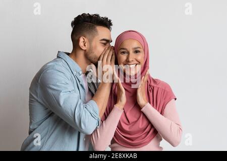 Klatsch-Konzept. Fröhlicher arabischer Kerl, der seiner muslimischen Freundin etwas ins Ohr flüstert Stockfoto