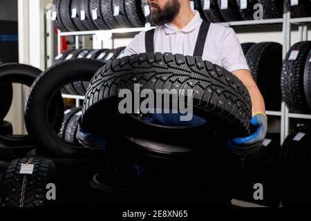 Automechaniker in einer Werkstatt auf einem Reifenstapel an seinem Arbeitsplatz, junger Mann in Uniform hält Reifen in Händen Stockfoto