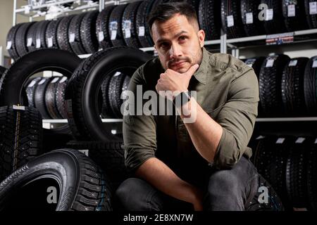 Männliche Kunden sitzen denken, welche der Reifen zu kaufen, kaukasischen Kerl in Freizeitkleidung sitzt in Kontemplation in Auto-Service-Shop, umgeben von schwarzen Reifen f Stockfoto