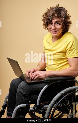 Behinderte Person in Casual Wear sitzend mit Laptop auf Rollstuhl, isoliert über beige Studio-Hintergrund. Gut aussehende lockig männlich genießen die Arbeit online, Typ Stockfoto