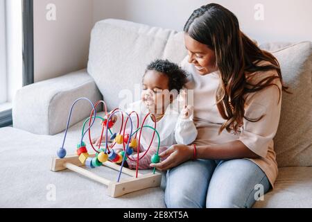 Glücklich lächelnd gemischte Rasse indische Mutter und afrikanische schwarz Baby Kleinkind spielen Entwicklung Holzperle Labyrinth Spielzeug zusammen zu Hause. Authentischer ehrlicher li Stockfoto