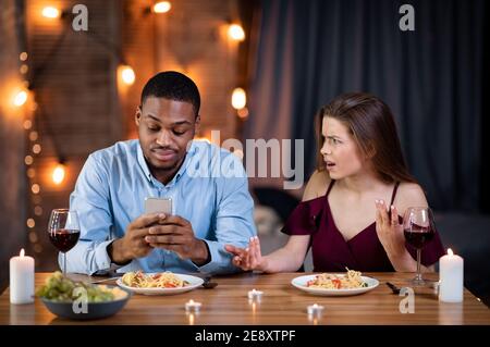 Phubbing-Konzept. Schwarzer Mann Beschäftigt Mit Smartphone Während Des Abendessens Mit Seiner Freundin Stockfoto
