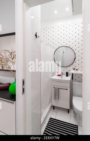 Modernes gefliestes, helles weiß-schwarzes Badezimmer und Toilette in einem kleinen Studio-Apartment. Innenansicht mit Schrank und Waschbecken, runder Spiegel, Dusche Stockfoto