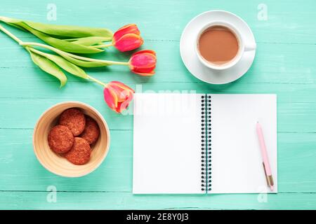 Leerer Notizblock, Kaffeetasse, Tulpen und Kekse auf türkisfarbenem Holztisch. Gemütlicher Arbeitsplatz, Frühlingsferien Konzept. Draufsicht, flach liegend, Kopierbereich. Stockfoto