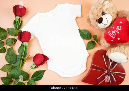 Happy Valentine's Day Baby Kleidung Flatlay owith Teddybär, rote Rosen und herzförmigen Geschenk auf modernen Korallen Hintergrund. Modell mit negativer Kopie sp Stockfoto