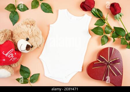 Happy Valentine's Day Baby Kleidung Flatlay owith Teddybär, rote Rosen und herzförmigen Geschenk auf modernen Korallen Hintergrund. Modell mit negativer Kopie sp Stockfoto