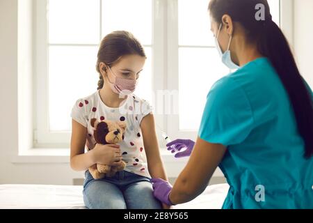 Doktor ist im Begriff, einen Impfstoff in die Hand eines niedlichen kleinen Mädchens zu injizieren, das ihr Lieblingsspielzeug hält. Stockfoto