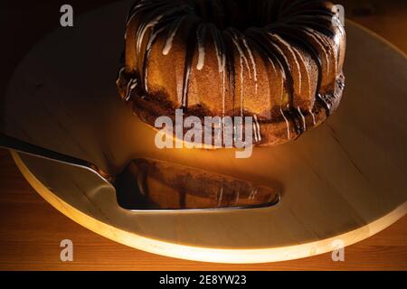 Brauner, zuckerbeschichteter Brundtkuchen auf einem weißen Drehteller mit silberner Kuchenschaufel, Bild auf der Vorderseite des Kuchens und der Schaufel fokussiert, warmes Licht Stockfoto