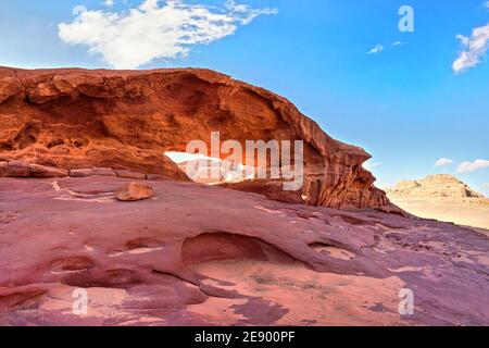 Kleiner Bogen oder kleine Felsenfensterformation in der Wadi Rum Wüste, helle Sonne scheint auf rotem Staub und Felsen, blauer Himmel darüber Stockfoto