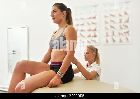 Physiotherapeutin trainiert mit ihrer Patientin, untersucht ihre unteren Rückenmuskeln, während sie vor ihr sitzt Stockfoto