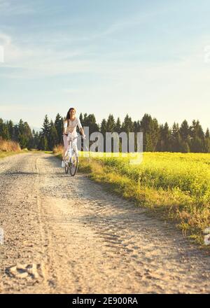 Junge Frau im Sommer leichte Kleidung fährt Fahrrad auf staubigen Straße in Richtung Kamera, Nachmittag Sonne scheint auf Felder und Wald im Hintergrund Stockfoto