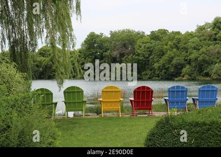 Sechs bunte Holzstühle an einem Seeufer umgeben von Wald in Janesville, Wisconsin, USA Stockfoto