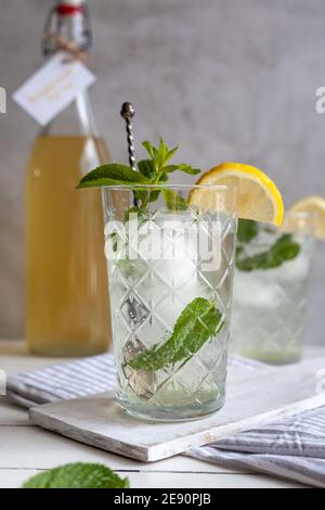 Erfrischendes Limonadengetränk mit Eis, Zitrone und Minze mit Sirup Flasche im Hintergrund, helle Oberfläche Stockfoto