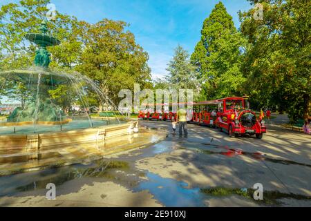 Genf, Schweiz - 15. Aug 2020: Brunnen der vier Jahreszeiten im Zentrum von Jardin Anglais mit rotem Touristenzug mit Waggons und Lokomotive Stockfoto