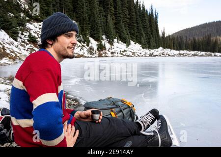 Seitenansicht des jungen Mannes in Pullover und Hut sitzend Am Ufer des gefrorenen Sees in der Nähe Nadelwald und Trinken Heißes Getränk während der Pause Stockfoto