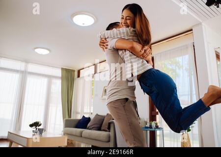 30s junge Erwachsene asiatische Mann hält Frau im Wohnzimmer zu Hause. Glückliches Paar dreht sich und dreht sich mit Aufregung in gemütlichen Haus. Happy Relationship Life Konzept. Stockfoto