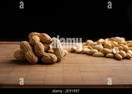 Geschälte und ungeschälte Erdnusskerne und auf Bambus-Schneidetisch mit schwarzem Hintergrund. (arachis hypogaea) essbare Samen. Gesunde Snack Ernährung Konzept Stockfoto