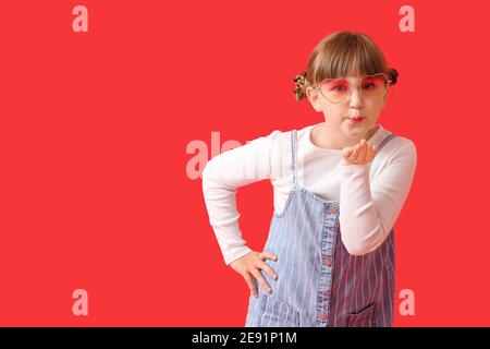 Nettes kleines Mädchen mit stilvollen Sonnenbrillen weht Kuss auf Farbe Hintergrund Stockfoto