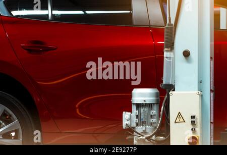 Selektiver Fokus auf rot glänzendes Auto und Pol der Aufzugsmaschine mit Motor und Motor-Steuerbox in der Autowerkstatt. Rotes SUV-Auto in der Garage angehoben. Stockfoto