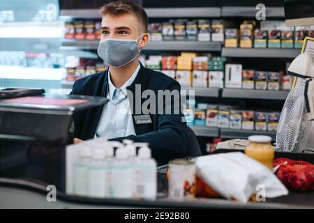 Männliche Trainee Kassierer in einem Lebensmittelgeschäft trägt eine Gesichtsmaske. Junge wesentliche Arbeiter arbeiten im Supermarkt während Pandemie. Stockfoto