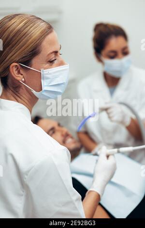 Nahaufnahme einer Zahnchirurgen-Frau, die eine Patientin in der Klinik mit ihrer Assistentin im Hintergrund behandelt. Zahnarzt, der einen männlichen Patienten in ihrem Büro behandelt. Stockfoto