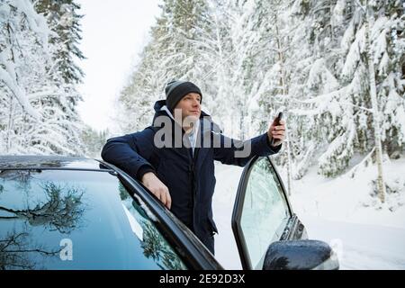 Mann in warmer Winterkleidung, der am Auto steht, mit dem Telefon. Verschneite Winter Landstraße, Auto mit Eis bedeckt, schöner Wald unter dem Schnee. Stockfoto