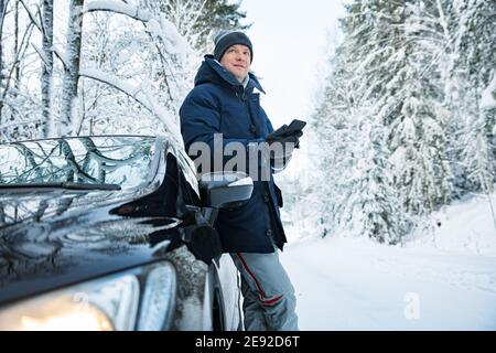 Mann in warmer Winterkleidung, der am Auto steht, mit dem Telefon. Verschneite Winter Landstraße, Auto mit Eis bedeckt, schöner Wald unter dem Schnee. Stockfoto