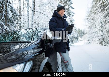 Mann in warmer Winterkleidung, der am Auto steht, mit dem Telefon. Verschneite Winter Landstraße, Auto mit Eis bedeckt, schöner Wald unter dem Schnee. Stockfoto