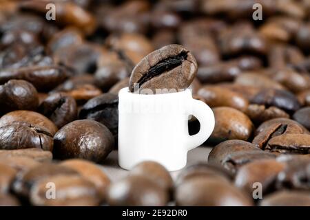 Miniatur weiße Kaffeetasse mit einer riesigen Kaffeebohne umgeben von verstreuten gerösteten Bohnen. Nahaufnahme, selektiver Fokus. Stockfoto