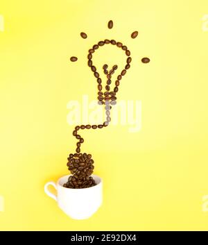 Kreative Anordnung von gerösteten Kaffeebohnen, die einer glühenden Glühbirne ähneln, die an eine weiße Kaffeetasse auf gelbem Hintergrund angeschlossen ist. Natürliche Energie booste Stockfoto