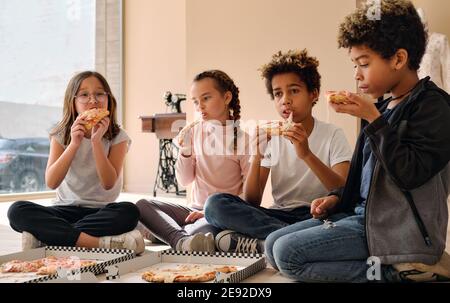 Gruppe von hungrigen multirassischen Kinder kleine Jungen Mädchen sitzen drinnen essen Pizza italienische Fast-Food-Küche. Alpha-Generierung teilt Slices, verbringt Zeit zusammen Stockfoto