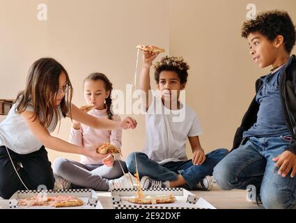 Gruppe von ziemlich multi ethnischen Kinder kleine Jungen Mädchen sitzen drinnen essen Pizza italienische Fast-Food-Küche. Alpha-Generierung teilt Slices, verbringt Zeit zusammen Stockfoto