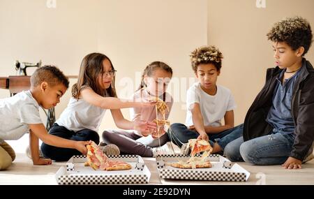 Fünf nette multirassische Kinder kleine Jungen und Mädchen sitzen drinnen essen Pizza italienische Fast-Food-Küche. Alpha-Generation teilen Slices, verbringen Zeit toge Stockfoto