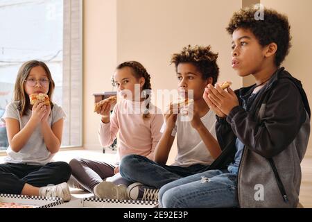 Gruppe von niedlichen multi Rasse Kinder kleine Jungen Mädchen sitzen drinnen essen Pizza italienische Fast-Food-Küche. Alpha-Generierung teilt Slices, verbringt Zeit miteinander Stockfoto