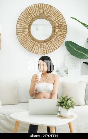 Junge asiatische schwangere Frau, die von zu Hause aus mit Computer und Trinken Milch für die gute Gesundheit ihres Babys Stockfoto