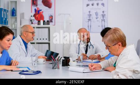 Medizinische Personal Leute auf der Personalversammlung sitzen vor einander am Tisch in weißen Mänteln und blauen Uniform-Shirts in einem Krankenhaus Büro, diskutiert medizinische Themen. Team von Ärzten Brainstorming. Stockfoto