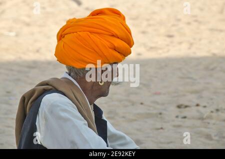 In der Morgensonne sitzt ein Kamelführer auf dem Messegelände. Der orangefarbene Turban auf seinem Kopf ist hell und attraktiv. Stockfoto