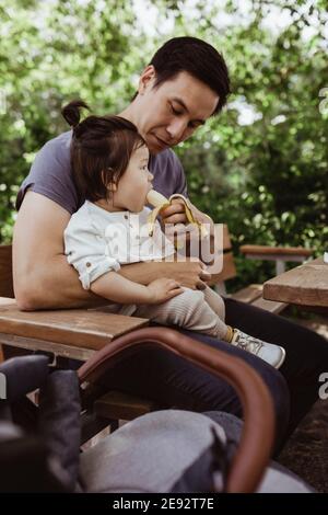 Vater Fütterung Banane zu männlichen Kleinkind, während im Park sitzen Stockfoto