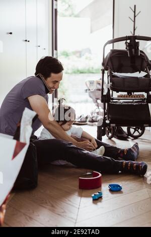 Vater im Gespräch auf Smartphone, während Baby Sohn Schnürsenkel binden Zu Hause Stockfoto