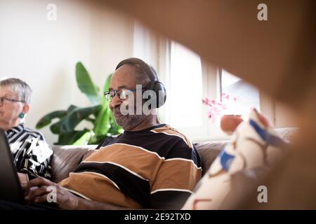 Lächelnder älterer Mann, der Musik über Kopfhörer hört, die von einer Frau gesessen werden Auf dem Sofa im Wohnzimmer Stockfoto