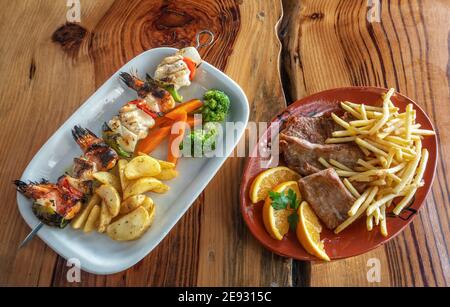 Gegrillter Fisch-Spieß mit Pommes und Gemüse auf der linken Seite. Teller Steak mit Pommes frites, garniert mit Orange auf der rechten Seite. Stockfoto